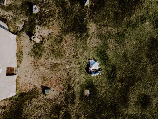 Fotograf fotí nevestu počas príprav pri okne, jemné prirodzené svetlo, svadobné šaty na vešiaku, emotívna realistická scéna bez textu