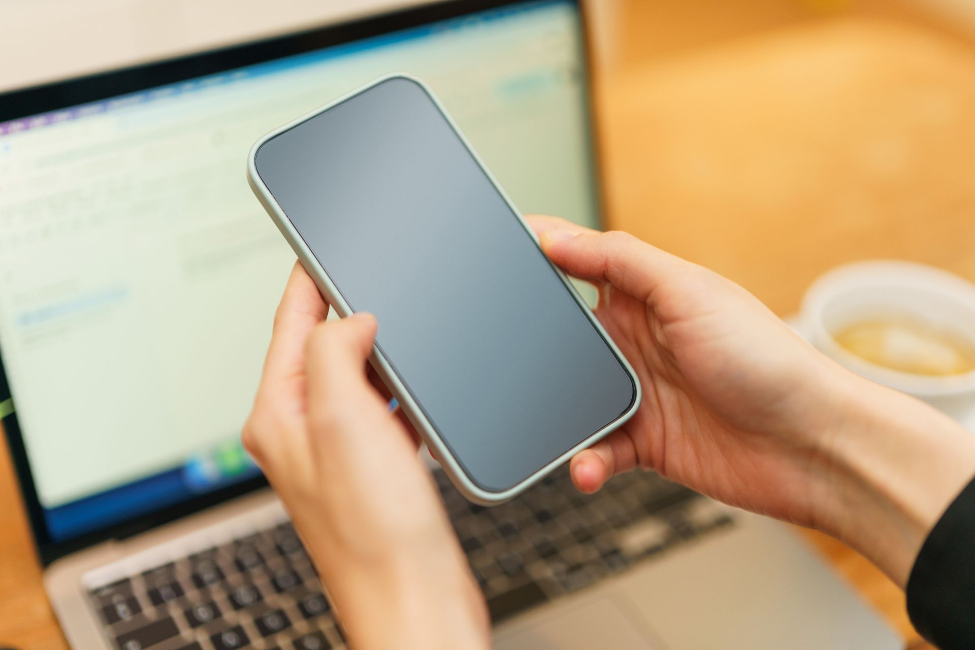 Ruky držiace mobilný telefón pri notebooku a šálke kávy